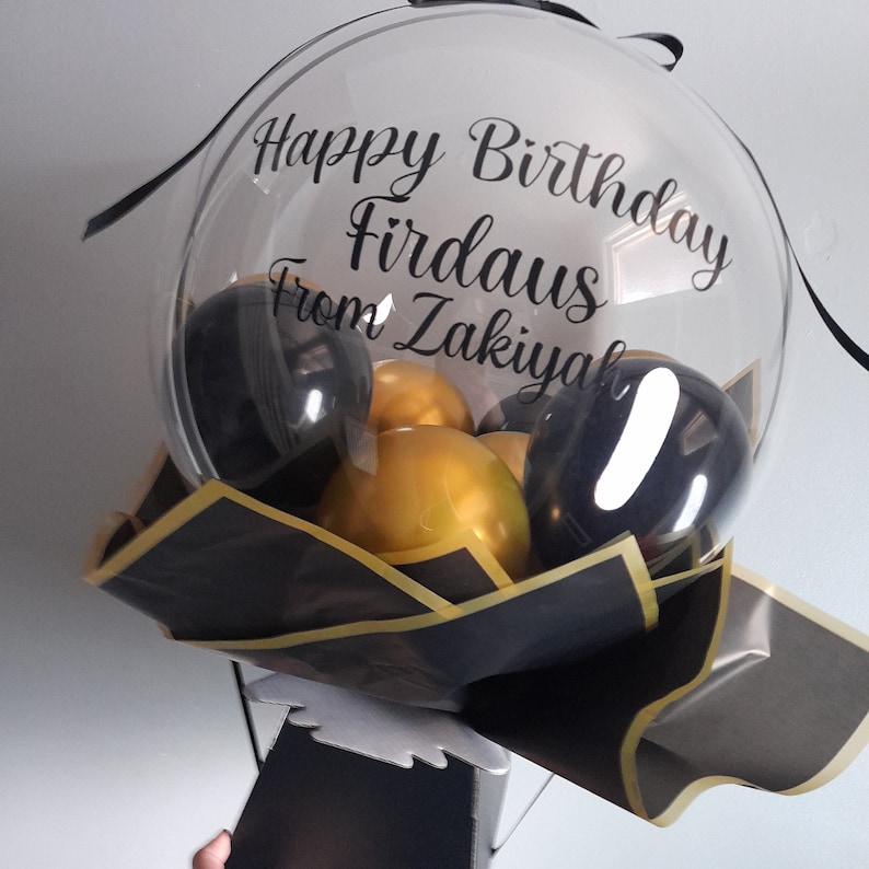 Pu&ograve; includere: Un bouquet di palloncini trasparente con palloncini neri e dorati. Il palloncino ha un messaggio che dice "Happy Birthday Firdaus from Zakiyah".