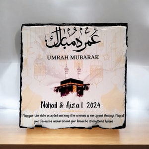 May include: A white and gold decorative slate plaque with black calligraphy that reads "Umrah Mubarak". The plaque features a watercolor illustration of the Kaaba, a mosque in Mecca, Saudi Arabia. The text "Nohail & Aiza I 2024" is printed below the illustration. The plaque includes a message wishing the couple a blessed Umrah pilgrimage.