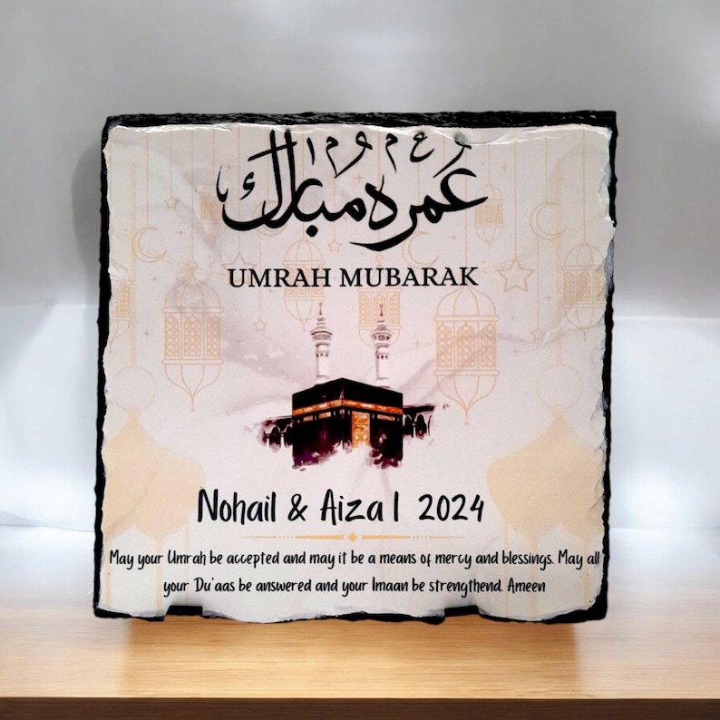 May include: A white and gold decorative slate plaque with black calligraphy that reads "Umrah Mubarak". The plaque features a watercolor illustration of the Kaaba, a mosque in Mecca, Saudi Arabia. The text "Nohail & Aiza I 2024" is printed below the illustration. The plaque includes a message wishing the couple a blessed Umrah pilgrimage.