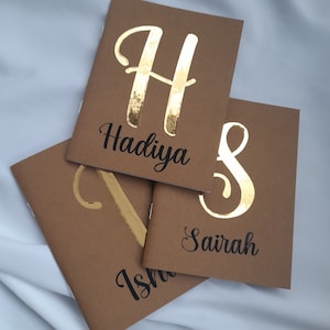 May include: Three brown notebooks with gold foil lettering. The notebooks are stacked on top of each other. The top notebook has the letter "H" and the name "Hadiya" written in gold foil. The middle notebook has the letter "S" and the name "Saira" written in gold foil. The bottom notebook has the letter "I" and the name "Isha" written in gold foil.