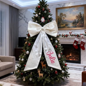 Può includere: Un albero di Natale decorato con un grande fiocco color crema e il nome "Julie" in corsivo rosa. L'albero è adornato con ornamenti rossi e dorati e decorazioni di renne in legno.