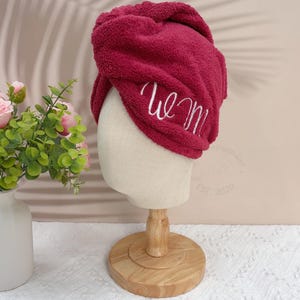 May include: A burgundy hair towel wrap with the embroidered word "we m" in white. The towel is displayed on a mannequin head with a wooden stand. The background includes a vase of pink roses.