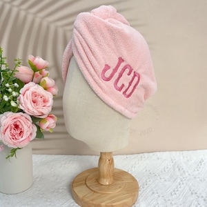May include: A light pink terry cloth hair towel with the embroidered initials "JCD" in a darker pink. The towel is displayed on a mannequin head with a wooden stand. Artificial pink roses in a white vase are in the background.
