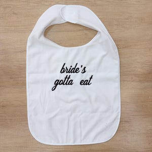 Può includere: Una bavaglina in tessuto bianco con scollo arrotondato e chiusura a strappo sulla parte superiore. Al centro della bavaglina è presente la scritta nera "bride's gotta eat" in carattere corsivo. La bavaglina ha un design semplice.