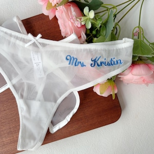 May include: White sheer thong underwear with a delicate bow and the embroidered text "Mrs. Kristin" in blue. The lingerie is displayed on a wooden surface with pink flowers in the background.