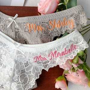 Könnte beinhalten: Nahaufnahme von zwei Spitzenunterhosen. Ein Paar ist weiß, das andere grau. Das weiße Paar hat den gestickten Text "Mrs. Mirabelle" in Rosa. Das graue Paar hat den gestickten Text "Mrs. Shirley" in Orange.