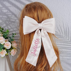 Op de afbeelding: Een grote, lichtroze satijnen haarstrik met lange linten. Het woord "Juliette" is in roze script op een lint geborduurd. De strik wordt in het haar gedragen, met de achterkant van het hoofd en lang, lichtbruin haar zichtbaar.