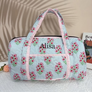 Könnte beinhalten: Hellblaue, gesteppte Reisetasche mit einem Blumenmuster in Rosa und Grün. Die Tasche hat einen schwarzen Reißverschluss, rosa und weiß gestreifte Paspeln und den Namen "Alisa" auf der Vorderseite gestickt. Die Tasche hat zwei Griffe.