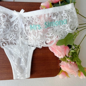 May include: White lace underwear with a delicate floral pattern and a small bow. The text "Mrs. Simona" is embroidered in light blue thread across the waistband. The garment is displayed with pink roses.