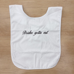 Puede incluir: Un babero blanco con escote redondeado y bordes rematados. El texto "Brides gotta eat" está impreso en letra cursiva negra en el centro del babero. La tela parece ser un material suave y absorbente con un diseño sencillo y limpio.
