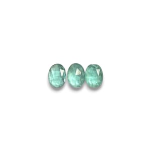 May include: Three oval-shaped, light teal gemstones are displayed against a white background. The gems have a faceted cut, reflecting light and showcasing their translucent quality. The gems are likely for jewelry making.