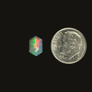 Single Ethiopian opal prism cut elongated hexagon gemstone cabochon.  Iridescent rose cut cabochon stone size 10 x 6.6 mm pictured on a black background next to a dime for size reference.