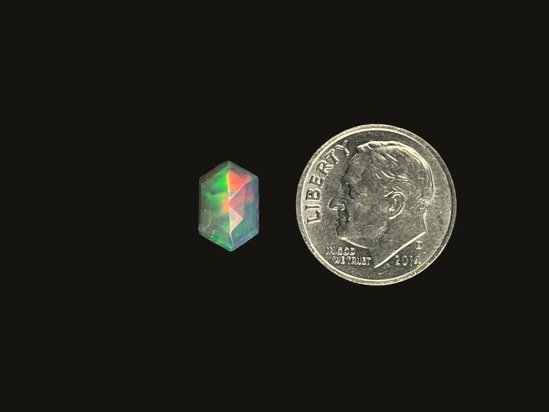 Single Ethiopian opal prism cut elongated hexagon gemstone cabochon.  Iridescent rose cut cabochon stone size 10 x 6.6 mm pictured on a black background next to a dime for size reference.