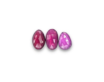 May include: Three faceted gemstones in shades of red and pink are displayed against a white background. The gemstones have an oval and teardrop shape. The gems are cut to reflect light, showcasing their color and clarity.