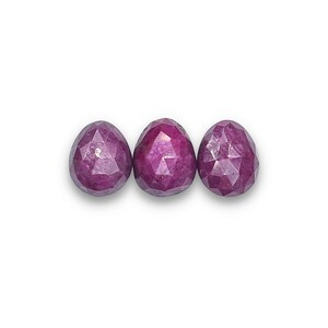 May include: Three faceted, oval-shaped gemstones in a deep magenta hue are arranged horizontally against a white background. The gemstones have a symmetrical, egg-like form, with multiple facets catching the light.