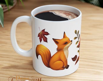 May include: A white ceramic mug filled with dark coffee, featuring a printed illustration of a small orange fox sitting next to a red maple leaf and a sprig of foliage. The text Dame Camomille is printed in a cursive font at the bottom.