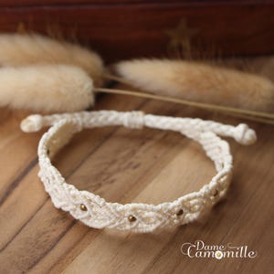 以下が含まれることがあります： 金色のビーズが付いた白いマクラメブレスレット。ブレスレットは結び目の技術で作られており、繊細でボヘミアンなスタイルです。
