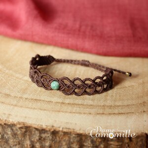 May include: A brown macrame bracelet with a green gemstone bead. The bracelet is on a wooden surface.