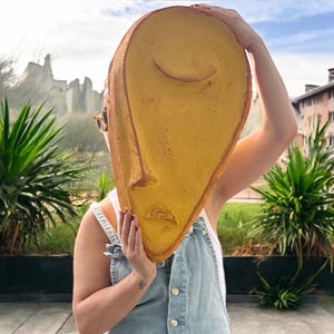 Puede incluir: Una persona sostiene una gran máscara facial de cerámica amarilla frente a su cara. La máscara tiene un diseño simple y estilizado con los ojos cerrados y una boca ligeramente hacia abajo.