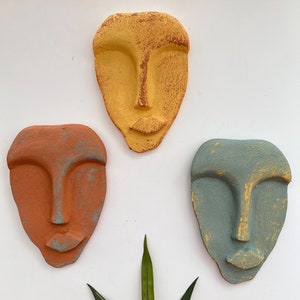 Puede incluir: Tres esculturas abstractas de rostros hechas de yeso o arcilla. Los rostros están pintados en tonos de amarillo, naranja y azul. Las esculturas se muestran sobre un fondo blanco.