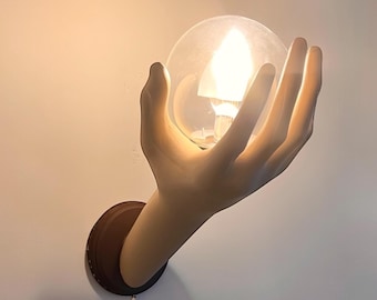 Puede incluir: Una escultura de mano de cerámica blanca sosteniendo una lámpara de globo de vidrio transparente con una bombilla de luz blanca cálida.