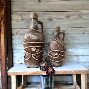 May include: Two brown ceramic bottles with rope accents and a decorative design. The bottles are sitting on a wooden shelf with a white surface.