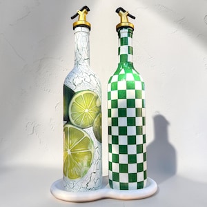 Könnte beinhalten: Zwei dekorative Glasflaschen mit goldfarbenen Ausgüssen und einem weißen Keramiktablett. Eine Flasche zeigt ein Limettendesign, die andere ein grün-weißes Schachbrettmuster. Die Flaschen sind für Öl oder Essig bestimmt.