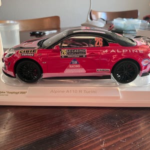 Könnte beinhalten: Ein rotes Alpine A110 R Turini Modellauto mit schwarzen Rädern und Rennaufklebern. Das Auto hat die Nummer 26, Sponsorenlogos und das Wort "ALPINE" an der Seite. Das Modell wird in einer transparenten Hülle präsentiert.