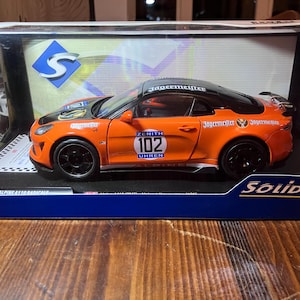 Könnte beinhalten: Ein orangefarbenes und schwarzes Modellauto mit der Nummer 102 auf der Tür. Das Auto trägt das "Jägermeister"-Logo und wird in einer Vitrine präsentiert. Das Modell ist ein Solido Alpine A110 Radical.