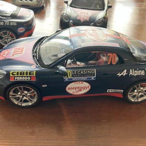 Könnte beinhalten: Ein schwarzes und rotes Spielzeug-Rennauto mit der Nummer 70 an der Seite und auf dem Dach. Das Auto hat Sponsorenlogos, darunter "CIBIE" und "FERODO". Der Text "4 Alpine 70 ans" ist ebenfalls sichtbar. Das Auto hat silberne Räder.