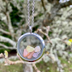 May include: A silver chain necklace with a round glass locket pendant containing small colorful stones. The locket is open and the stones are visible.
