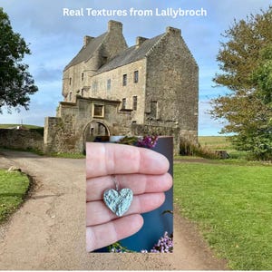 Puede incluir: Un colgante plateado en forma de corazón con una superficie texturizada se sostiene frente a un castillo de piedra. El colgante está en un gancho plateado. El fondo incluye un castillo de piedra, un camino y hierba verde. El texto "Real Textures from Lallybroch" está en la parte superior.