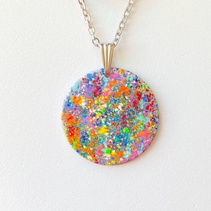 May include: A round pendant necklace with a silver chain. The pendant is a colorful mix of paint splatters in shades of red, orange, yellow, green, blue, and purple. The pendant is approximately 1.5 inches in diameter.