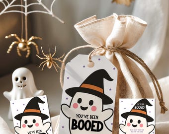 May include: Halloween-themed gift bag with a tag that says "You've Been Booed" and a cute ghost illustration wearing a witch hat. The image also includes a small ghost figurine, spiders, and a bat, all in a festive Halloween setting.