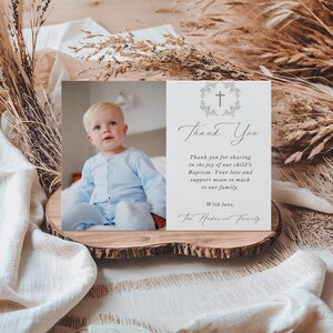 May include: A baptism thank you card with a photo of a baby in a blue outfit. The card features the text "Thank You" and a message of gratitude, along with a cross and floral design. The card is displayed on a wooden slice.