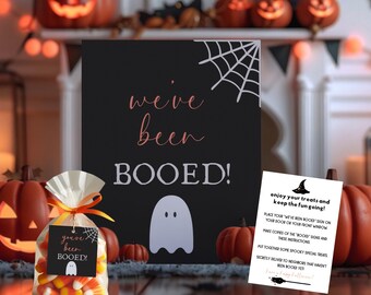 May include: A black Halloween sign with the words "we've been BOOED!" in white and pink script, a ghost graphic, and a spiderweb. A bag of candy corn and a small instruction card are also visible.