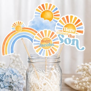 Op de afbeelding: Foto-rekwisieten voor een babyshower, waaronder een regenboog, zonnen en een wolk. De rekwisieten zijn in oranje, geel en blauw, met de tekst "Baby Jacobs" en "Here comes the Son". De rekwisieten zitten op houten stokjes.