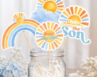May include: A collection of baby shower photo booth props featuring a rainbow, suns, and a cloud. The props are in shades of orange, yellow, and blue, with the text "Baby Jacobs" and "Here comes the Son". The props are on wooden sticks.