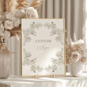May include: A framed sign with the words "CUSTOM Sign" in elegant script. The sign is surrounded by a floral design and set on a gold stand. The background features floral arrangements in vases.
