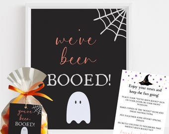 May include: A Halloween-themed image featuring a black sign with the words "we've been BOOED!" in white and a white ghost illustration. A bag of candy corn with a tag that says "you've been BOOED!" is in the foreground. A small instruction card is also present.