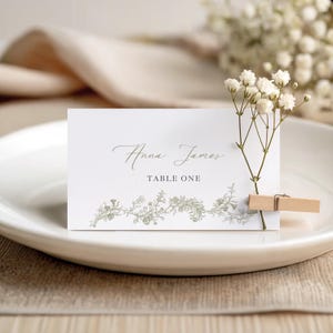 May include: A white place card with the names "Anna James" in elegant script and "TABLE ONE" printed below. The card features a delicate floral design and is held by a wooden clip. A sprig of white flowers is attached.
