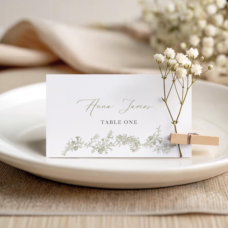 May include: A white place card with the names "Anna James" in elegant script and "TABLE ONE" printed below. The card features a delicate floral design and is held by a wooden clip. A sprig of white flowers is attached.