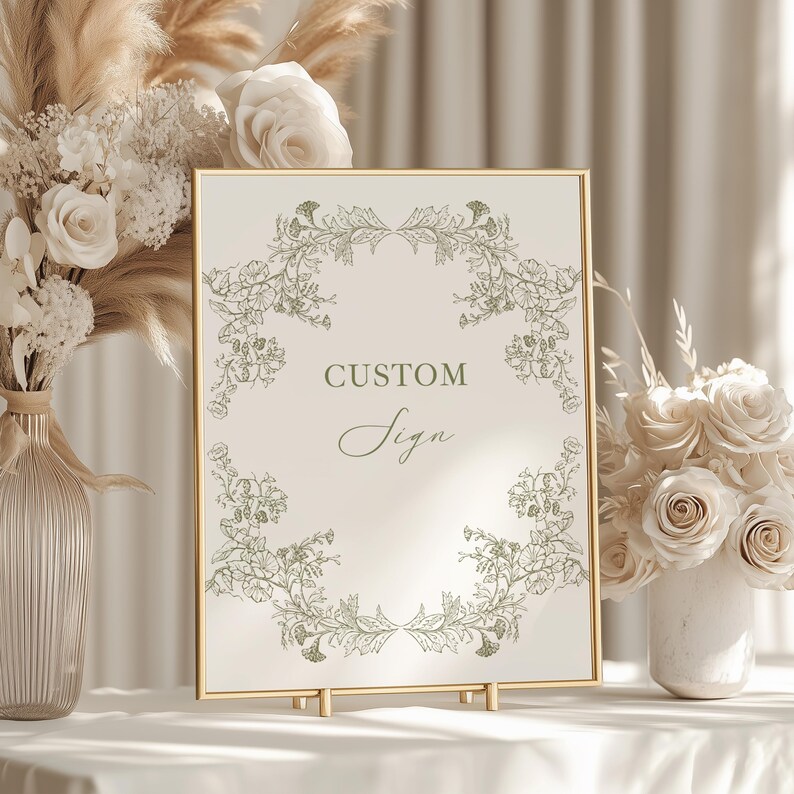 May include: A gold-framed sign displaying the words "CUSTOM Sign" in a decorative font. The sign features a floral border in green and gold tones. It is set on a white surface, with floral decorations in the background.