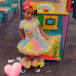 Pu&ograve; includere: Una bambina che indossa una maglietta bianca con una grafica di un personaggio di CoComelon, una gonna di tulle arcobaleno e scarpe colorate. &Egrave; in piedi davanti a una macchina da gioco con un adesivo a forma di cuore sul pavimento.