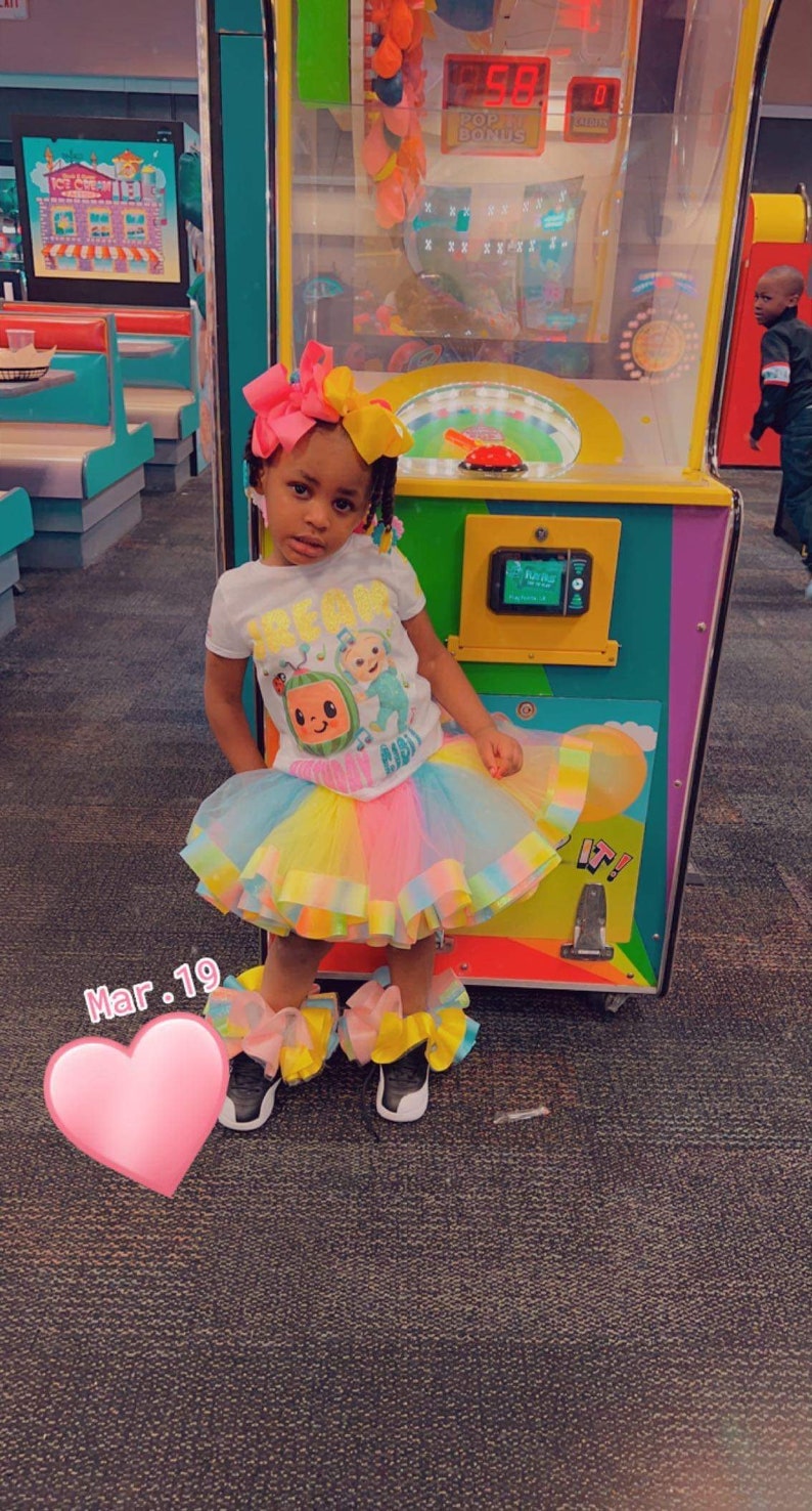 Pu&ograve; includere: Una bambina che indossa una maglietta bianca con una grafica di un personaggio di CoComelon, una gonna di tulle arcobaleno e scarpe colorate. &Egrave; in piedi davanti a una macchina da gioco con un adesivo a forma di cuore sul pavimento.