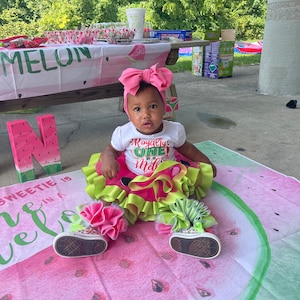 Pu&ograve; includere: Una bambina con una maglietta bianca a tema anguria, una fascia rosa e un tut&ugrave; verde e rosa &egrave; seduta su una coperta con motivo a anguria. La coperta ha le parole "Sweetie is in a melon" stampate.