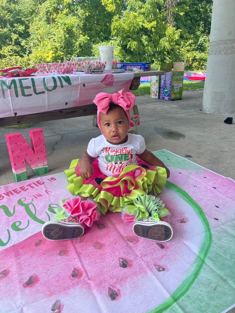 Pu&ograve; includere: Una bambina con una maglietta bianca a tema anguria, una fascia rosa e un tut&ugrave; verde e rosa &egrave; seduta su una coperta con motivo a anguria. La coperta ha le parole "Sweetie is in a melon" stampate.