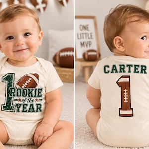 May include: A cream-colored baby bodysuit featuring a football-themed design. The front displays the text "1 ROOKIE of the YEAR" with a football graphic. The back shows the name "CARTER" above a large number "1" designed to look like a football.