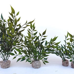 May include: A set of four artificial green plants with pointed leaves, each anchored in a small, circular, textured base covered in light tan gravel. The plants vary in height, arranged in a row from tallest to shortest against a white background.