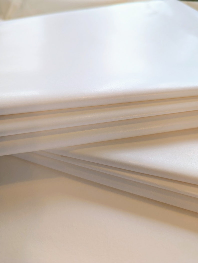 Puede incluir: Primer plano de una pila de hojas de papel de seda plegadas, de color blanquecino. El papel tiene una superficie lisa y ligeramente brillante. Los bordes del papel son visibles, mostrando las capas. La iluminaci&oacute;n crea sombras suaves, resaltando la textura y la forma del papel.
